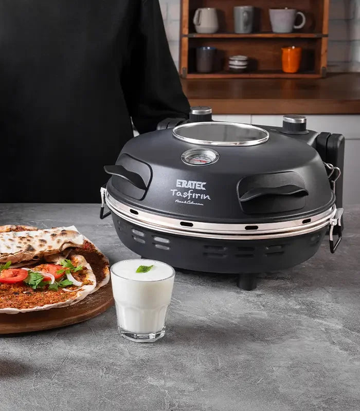 Schwarzer Steinofen auf Küchentisch mit Lahmacun und Glas Ayran daneben, Küchenregal im Hintergrund.