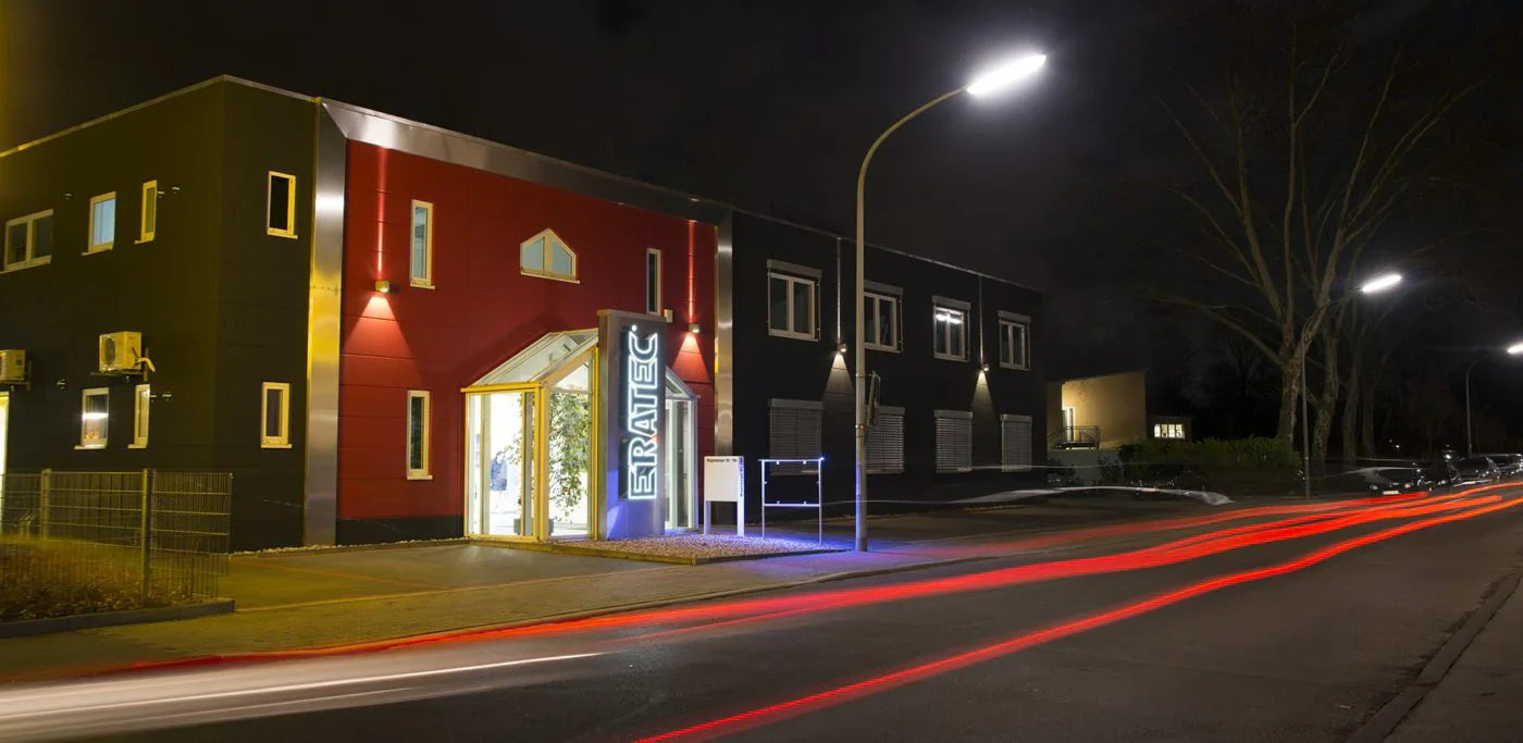 Modernes Bürogebäude bei Nacht mit beleuchtetem Eingang und Lichtspuren vorbeifahrender Autos.