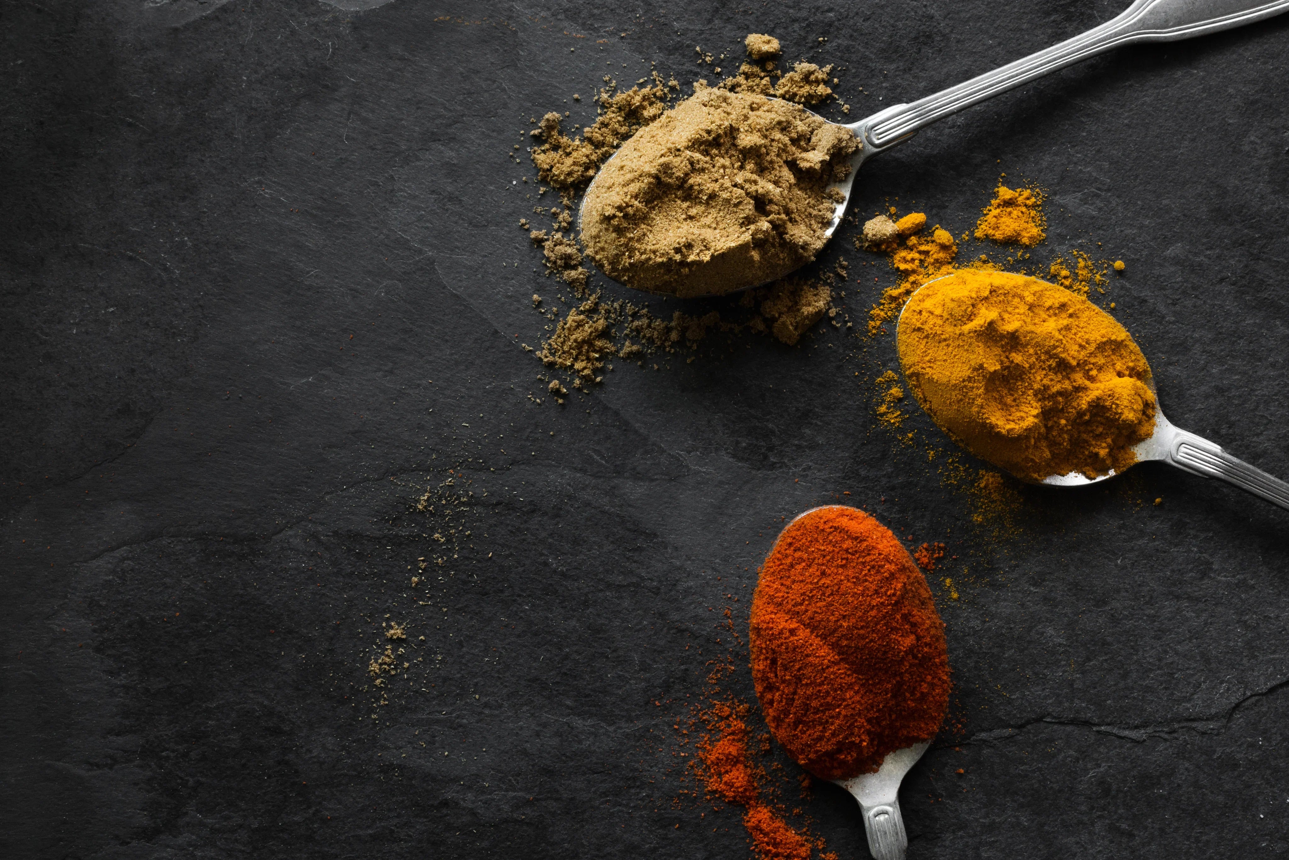 Drei Teelöffel mit bunten Gewürzen auf dunklem Stein, Paprika, Kurkuma, gemahlener Pfeffer